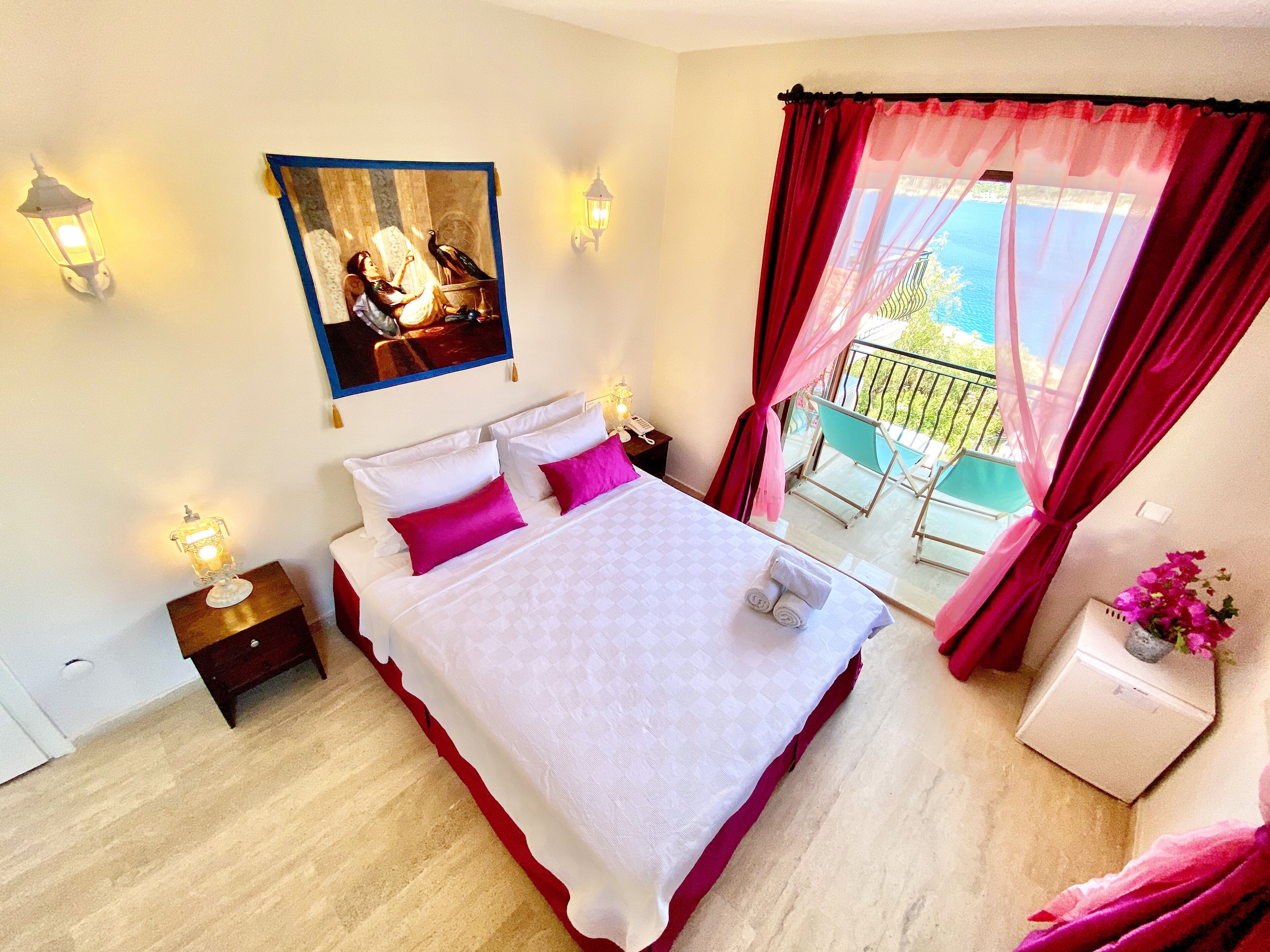 A hotel room with a bed, a painting, a balcony with a view, and a small table with a vase of flowers.
