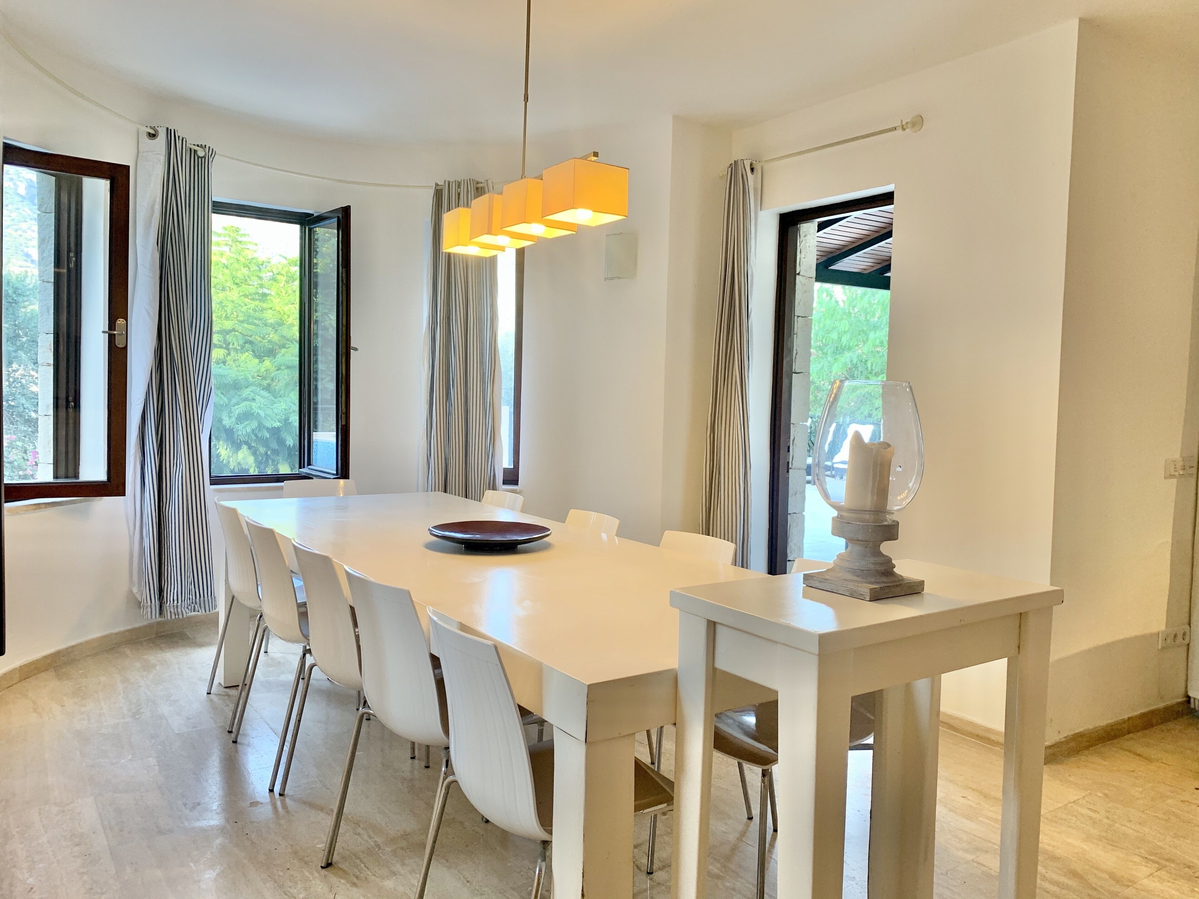 A modern dining area with a white table and chairs, a hanging light fixture, and large windows with curtains.