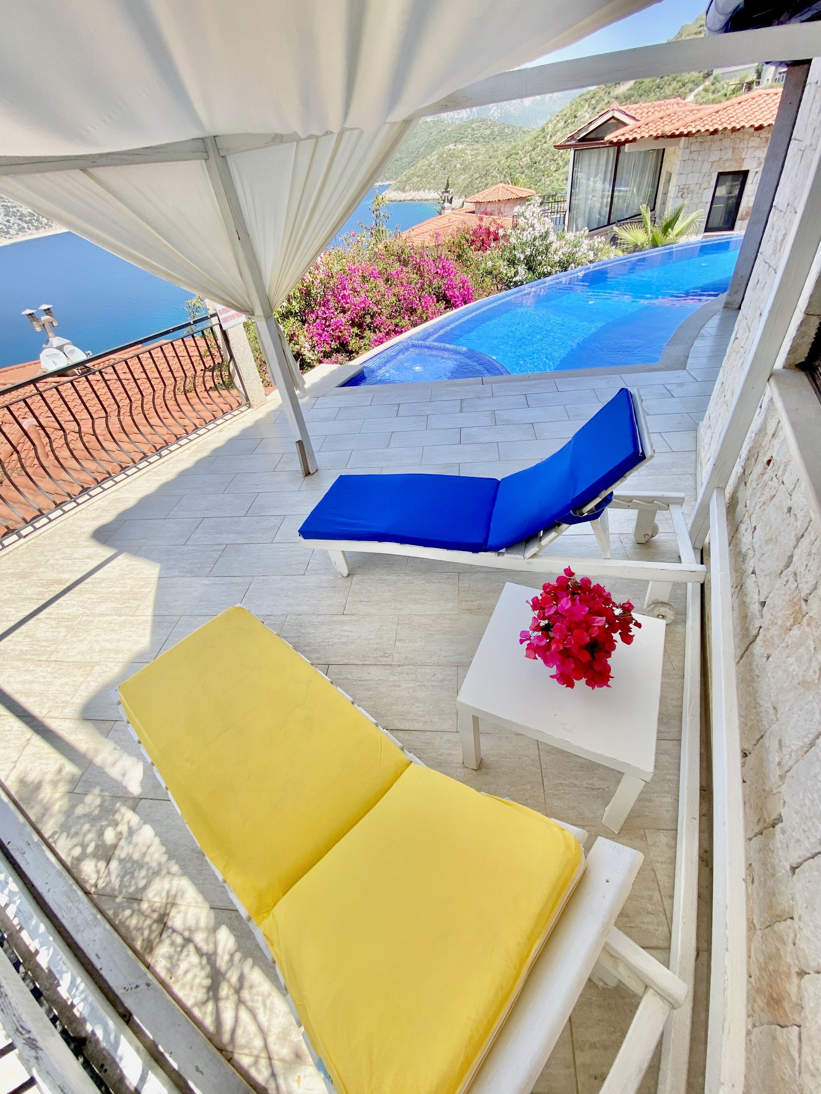 A terrace with two yellow lounge chairs, a blue lounge chair, a white table with a vase of pink flowers, and a view of a swimming pool and hillside buildings.