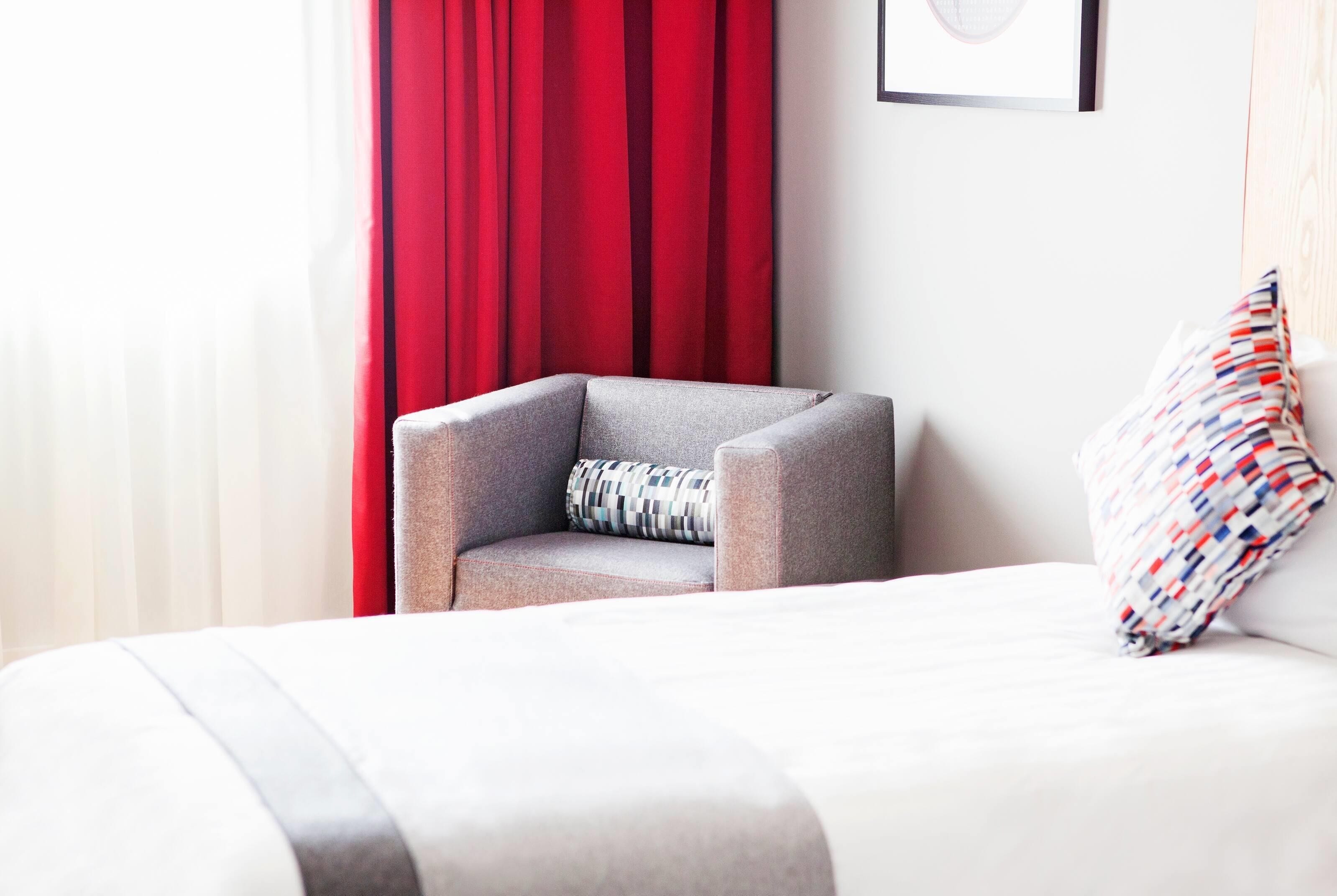 A modern hotel room with a grey armchair, a patterned pillow, and a bed with a striped blanket.