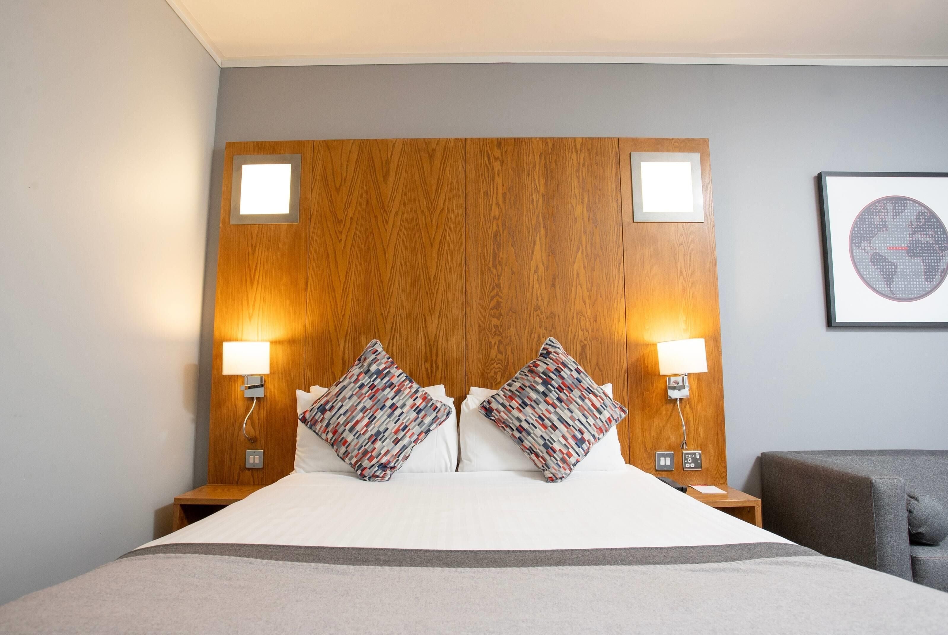 Une chambre d’hôtel avec une tête de lit en bois, deux lampes de chevet, un lit avec une literie blanche et un fauteuil gris.