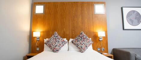 Ein Hotelzimmer mit einem hölzernen Kopfteil, zwei Nachttischlampen, einem Bett mit weißer Bettwäsche und einem grauen Sessel.
