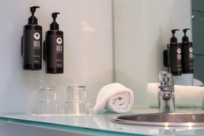 Un baño con un estante de vidrio que contiene dos botellas de jabón de manos, dos botellas de crema corporal y una toalla enrollada.