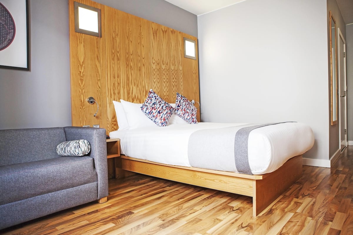 A modern bedroom with a wooden headboard, a grey sofa, and a bed with patterned pillows.
