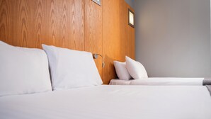 Una habitación de hotel con cabecera de madera, cama con ropa blanca y mesita de noche con lámpara.
