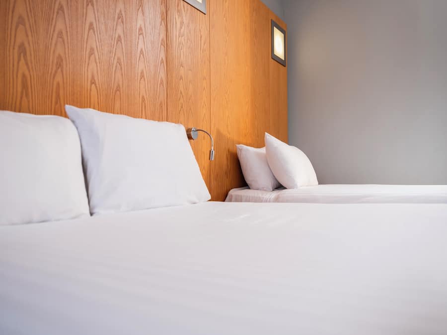 Quarto de hotel com cabeceira de madeira, cama com roupa de cama branca e mesa de cabeceira com abajur.