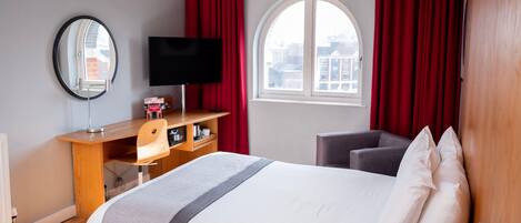 A hotel room with a bed, a desk with a computer, a television, and a window with curtains.