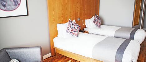 Cama doble con dos almohadas, cabecero de madera, sillón gris y mapa del mundo enmarcado en la pared.