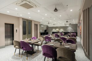 A modern dining area with multiple tables and chairs, a kitchen in the background, and a patterned floor. - Hotel Sunstar Grand (New Delhi)
