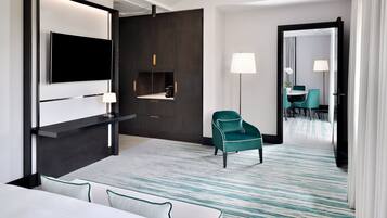 Ett modernt hotellrum med en platt-TV som är monterad på väggen, en sittgrupp med soffa och fåtöljer, ett litet bord samt en separat matplats med bord och stolar.