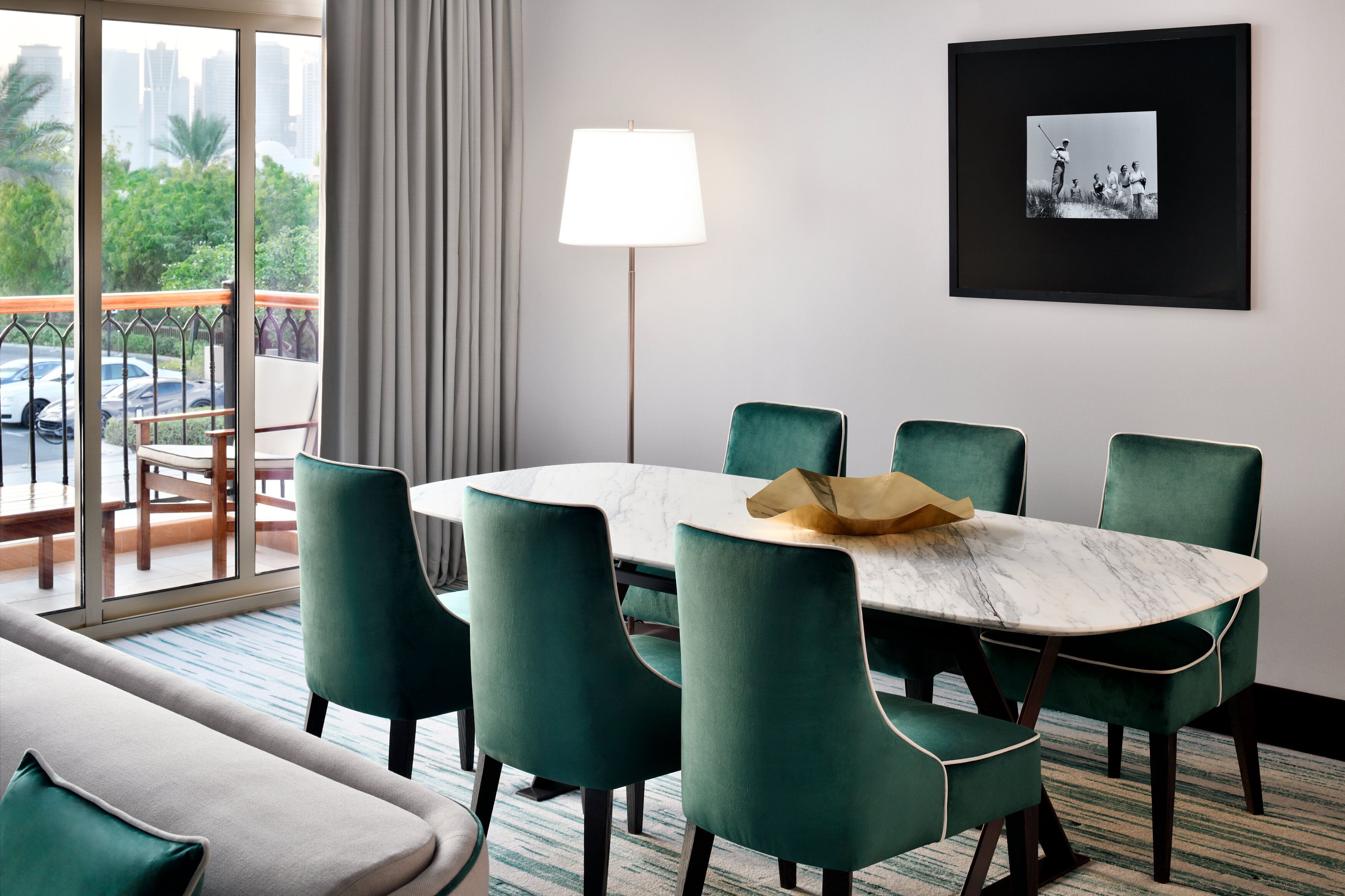 Ein Hotelzimmer mit einem runden Tisch, sechs grünen Stühlen, einer Stehlampe, einem gerahmten Foto an der Wand und einem Balkon mit Blick auf Palmen und Autos.