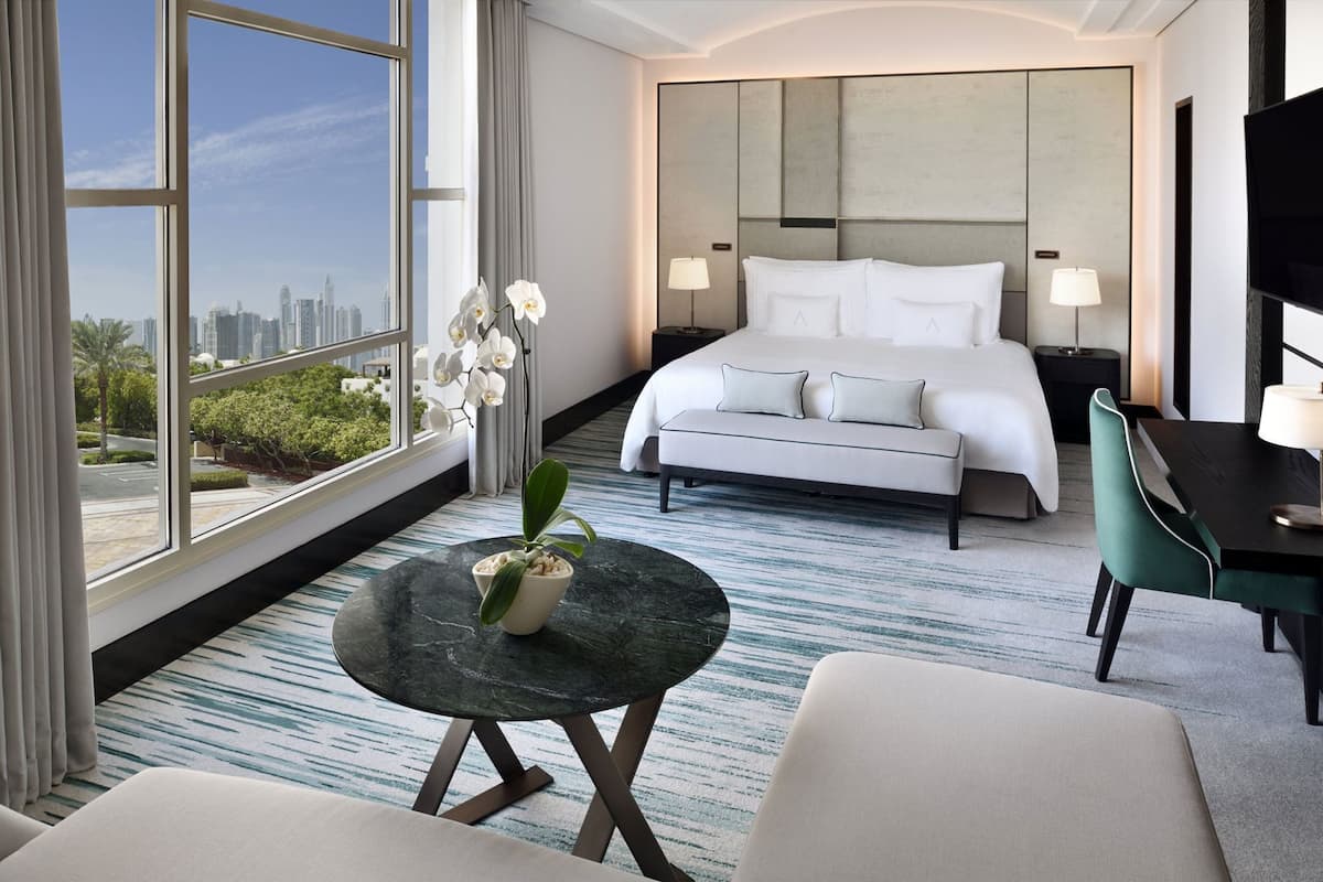 A hotel room with a bed, a desk, a chair, a small table with a plant, and a view of the city.