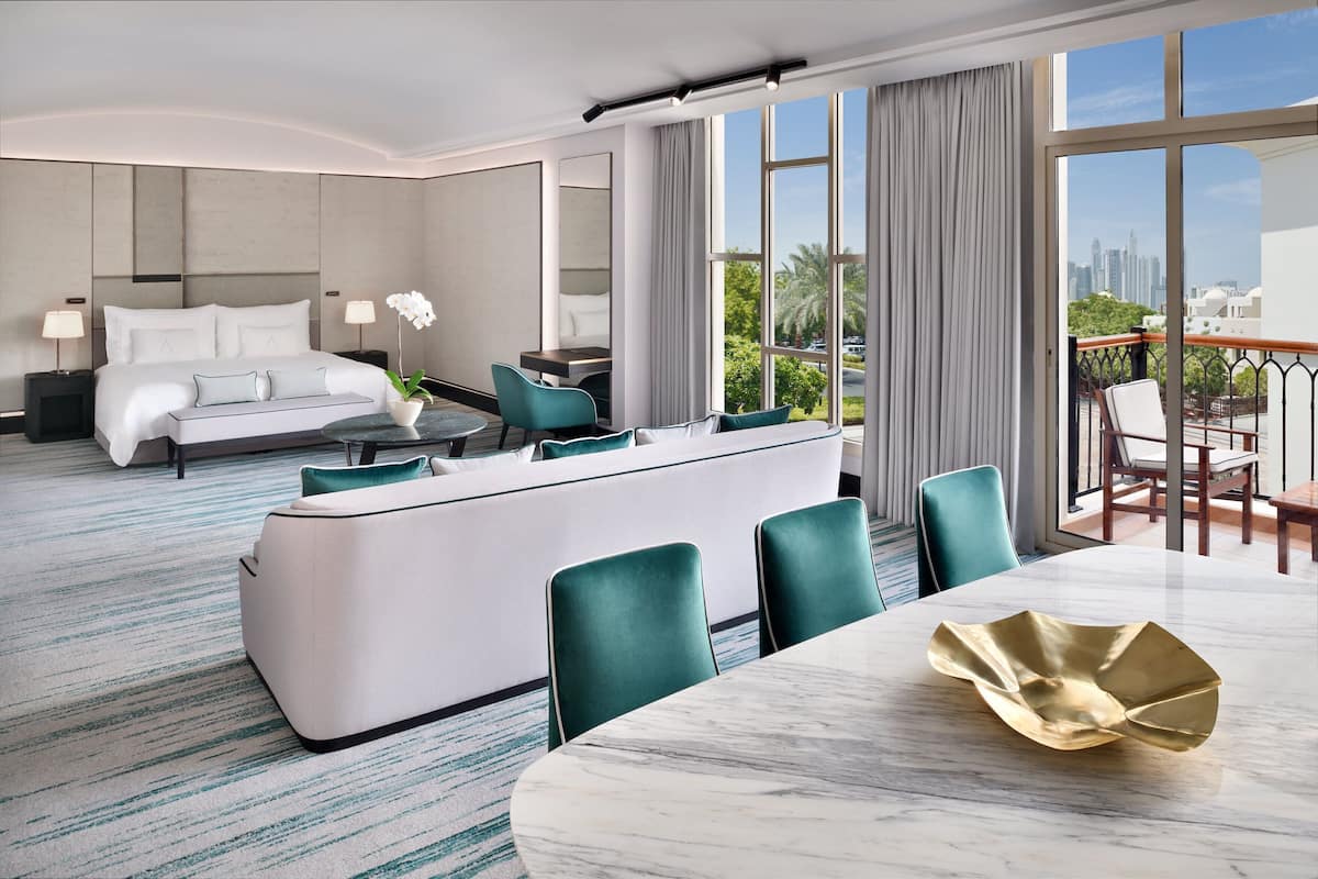 A modern hotel room with a bed, a sofa, a dining area, and a balcony with a city view.