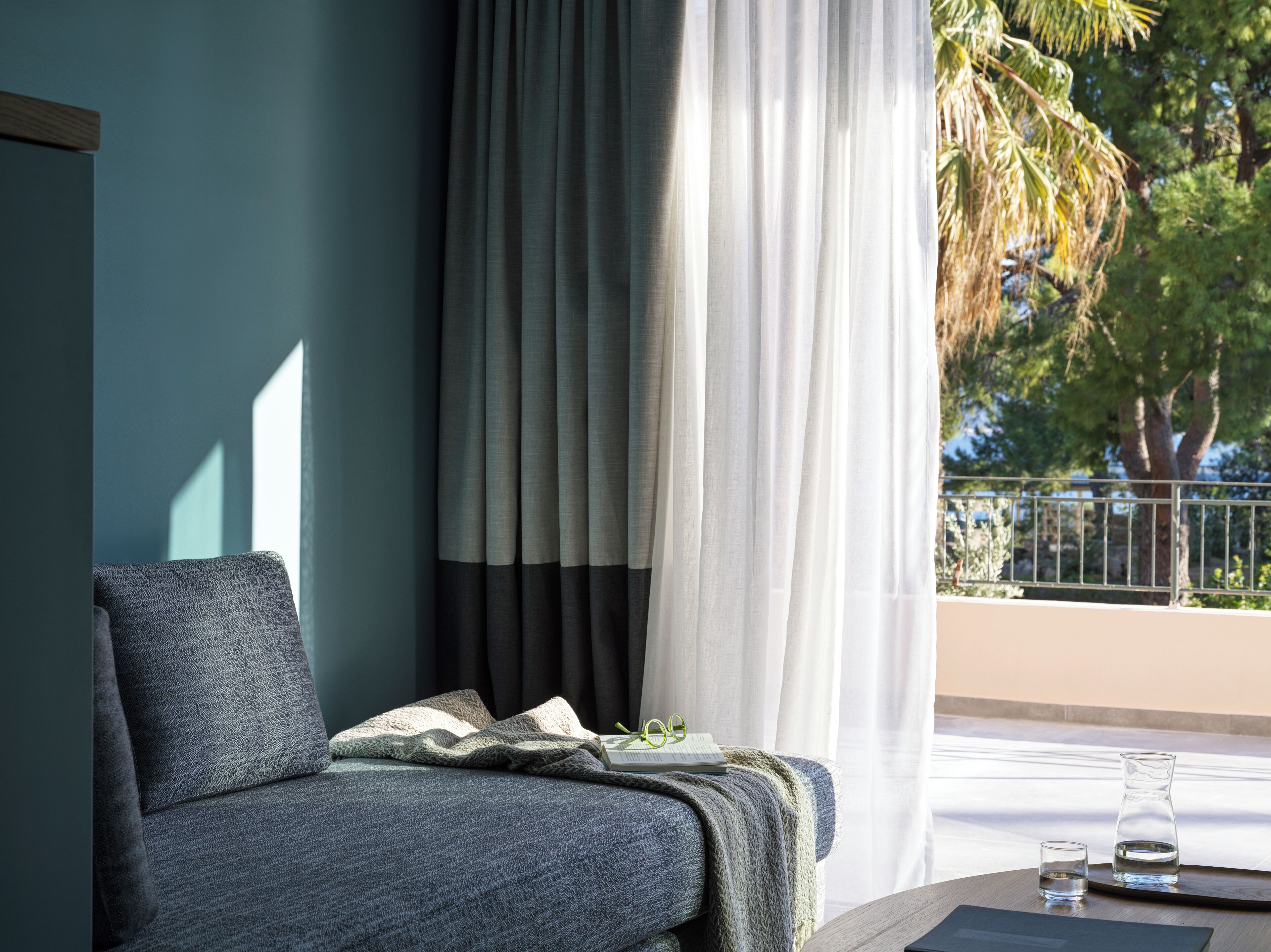 A modern room with a grey sofa, a small table with a glass of water, and a view of a balcony with a railing and trees.