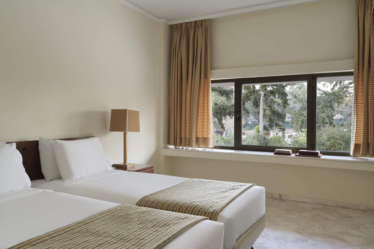 A hotel room with two beds, a window with curtains, a bedside table with a lamp, and a view of trees and houses outside.
