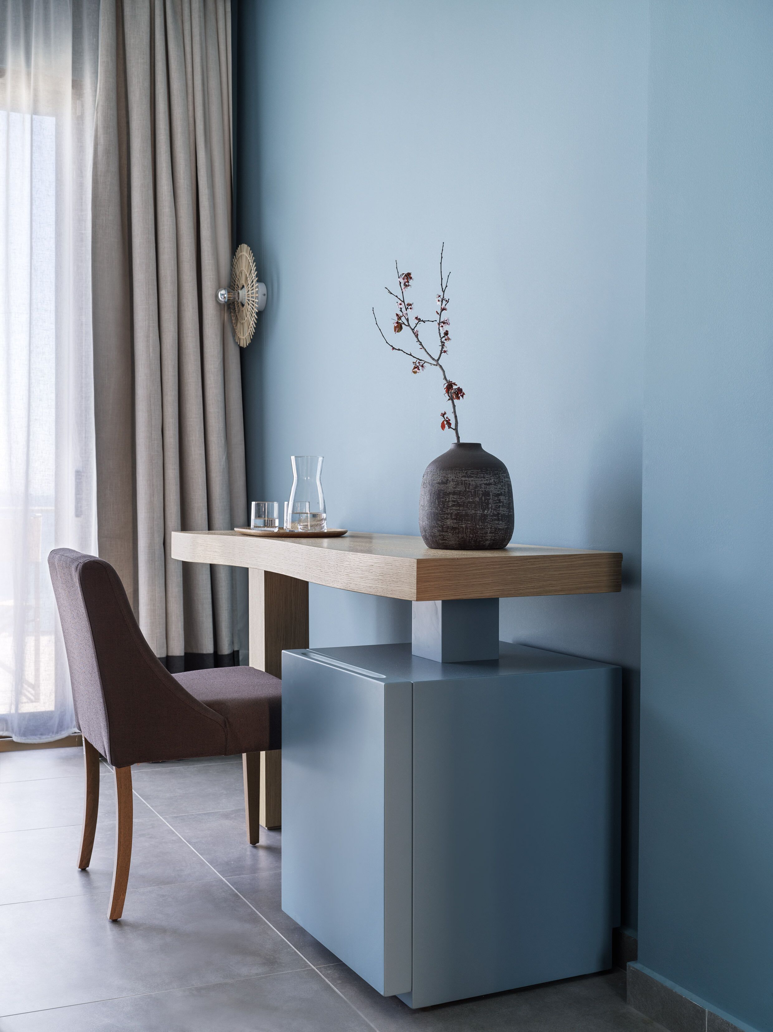 Ein modernes Zimmer mit minimalistischer Einrichtung, ausgestattet mit einem grauen Sofa, einem Holznachttisch und einer Vase mit einem Zweig.