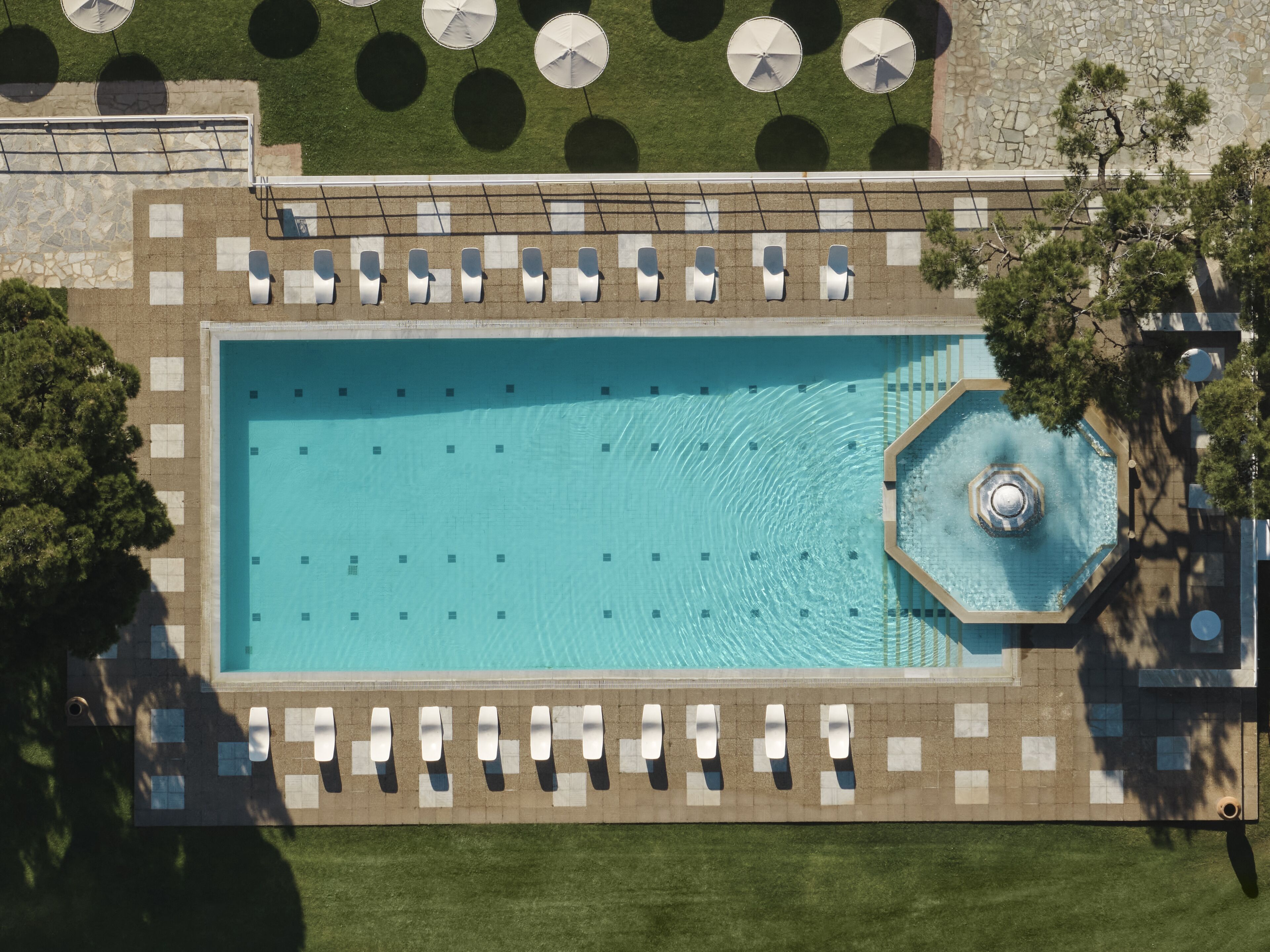 An aerial view of a rectangular swimming pool with a central fountain, surrounded by lounge chairs and umbrellas.
