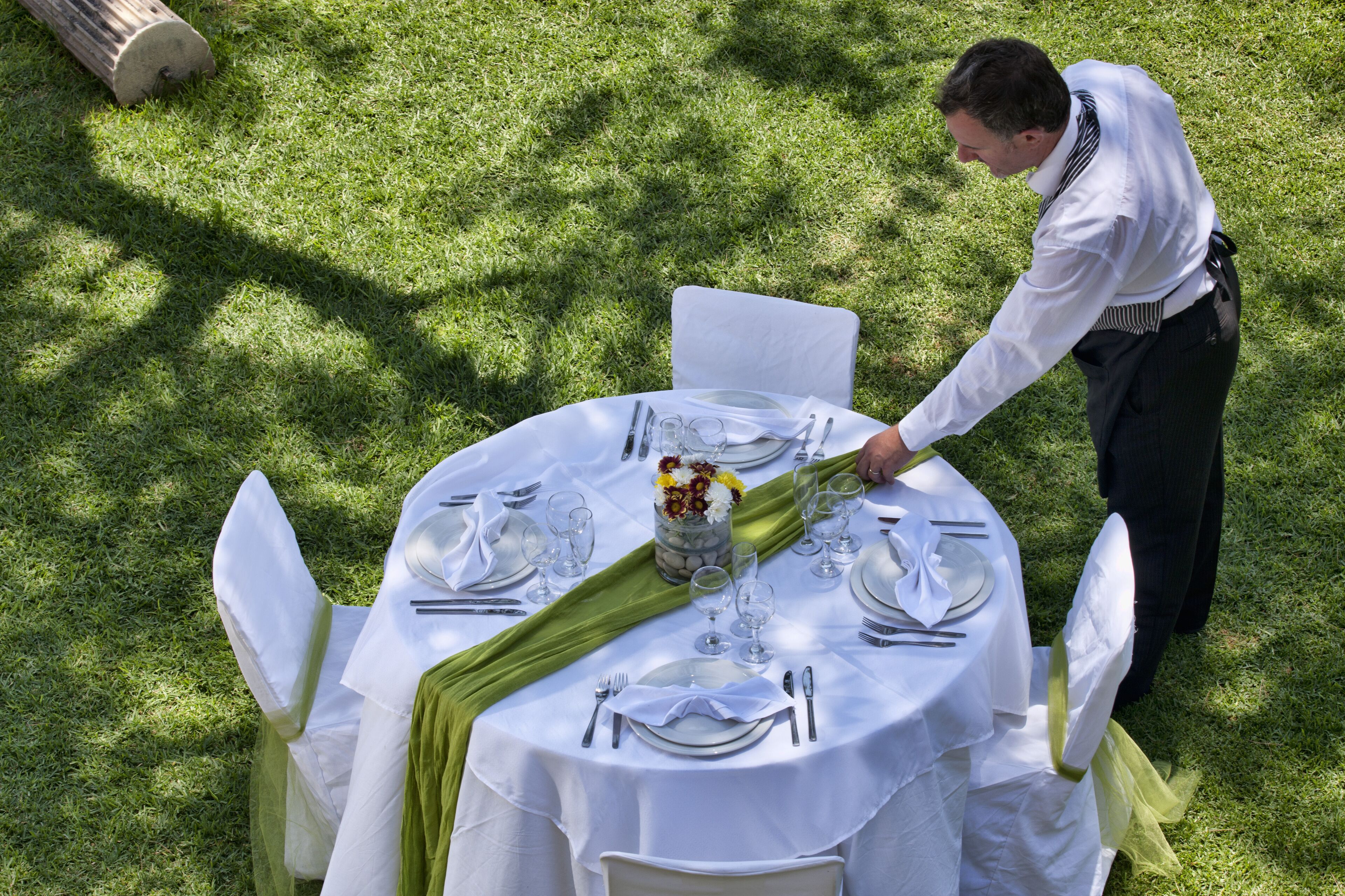 A round table set outdoors with white tablecloths, green napkins, and white chairs.