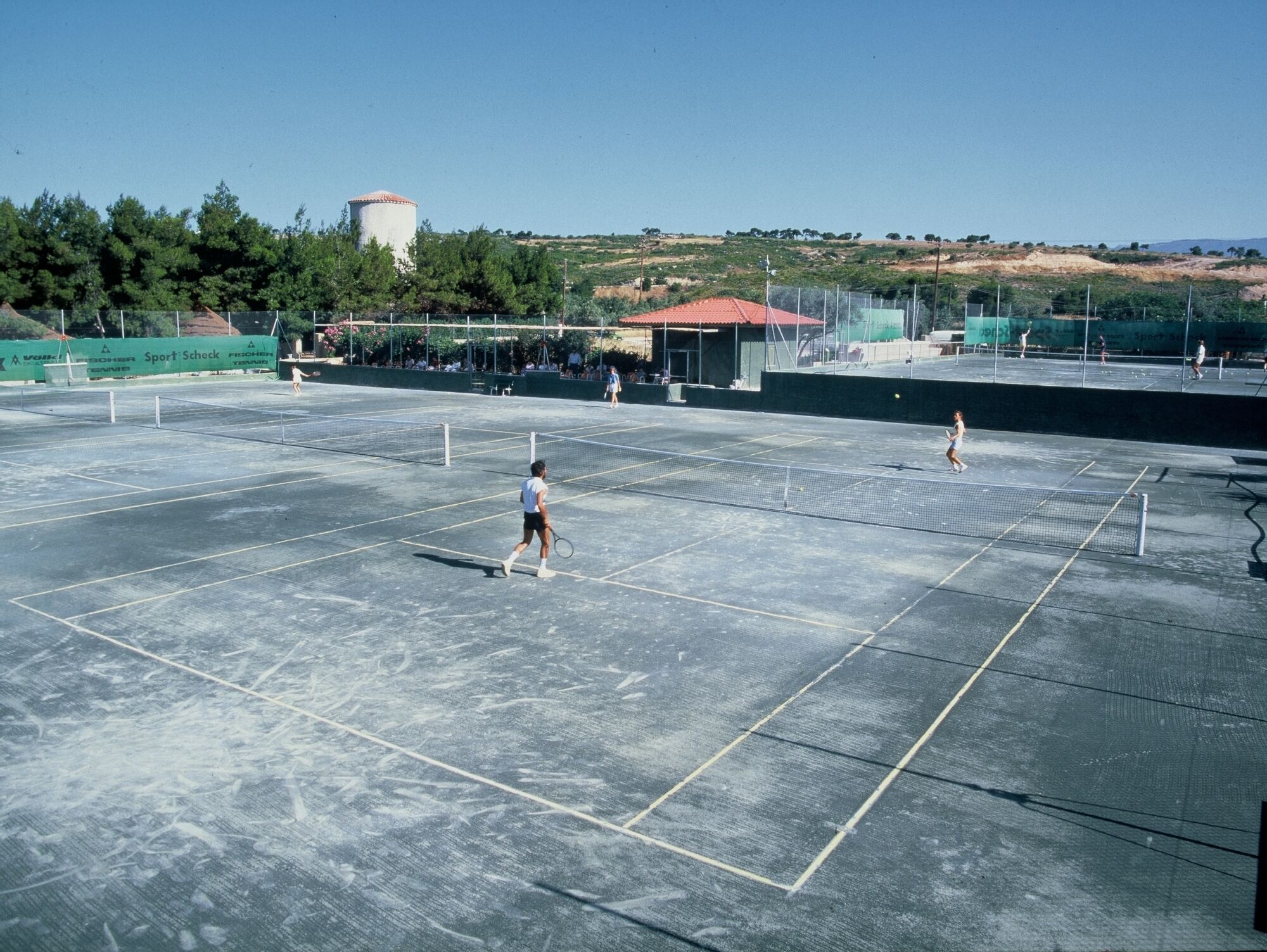 An outdoor tennis court with multiple courts, surrounded by trees and a hill in the background.