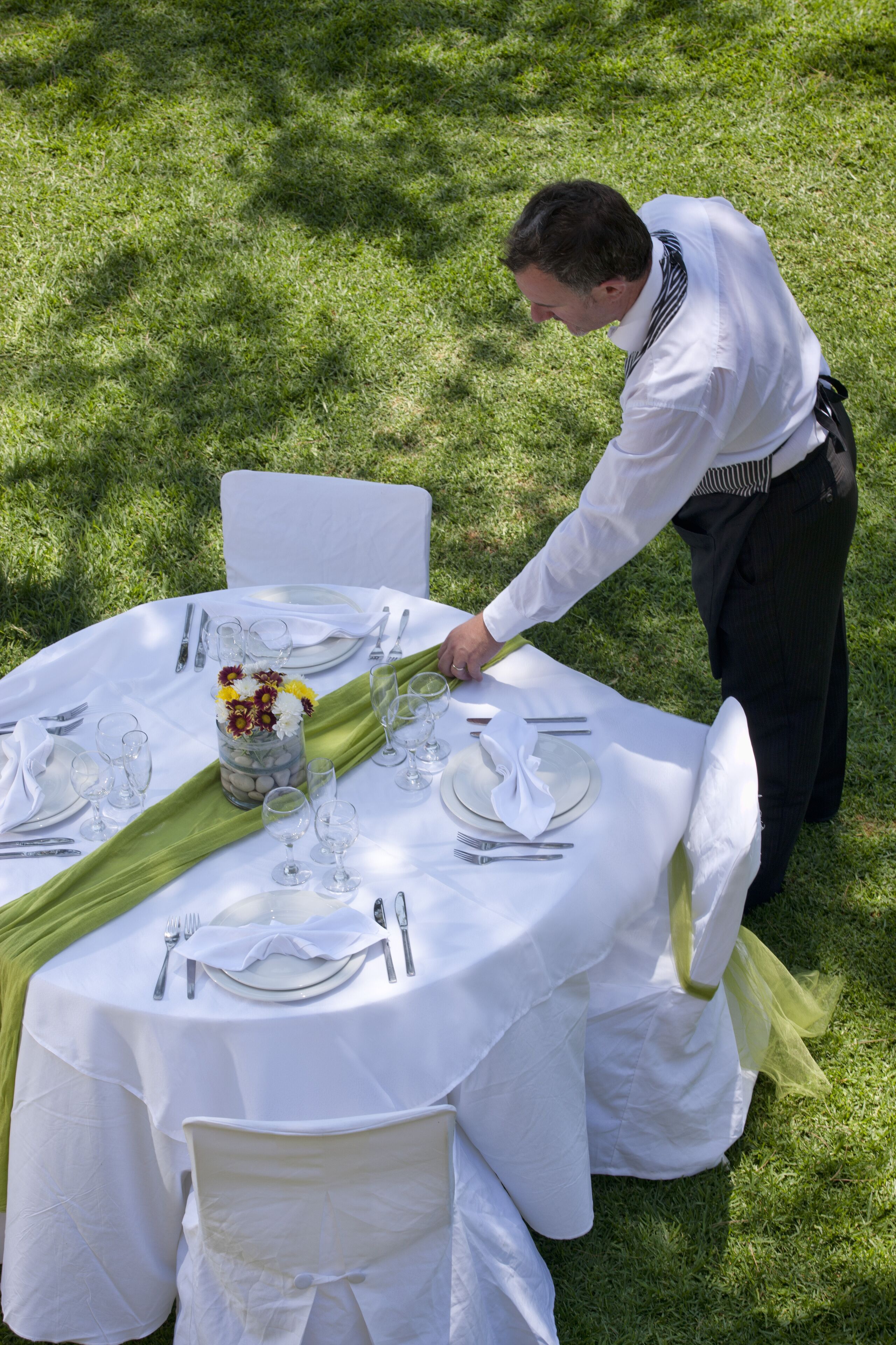 Una mesa redonda al aire libre con manteles blancos, servilletas verdes y un jarrón con flores.