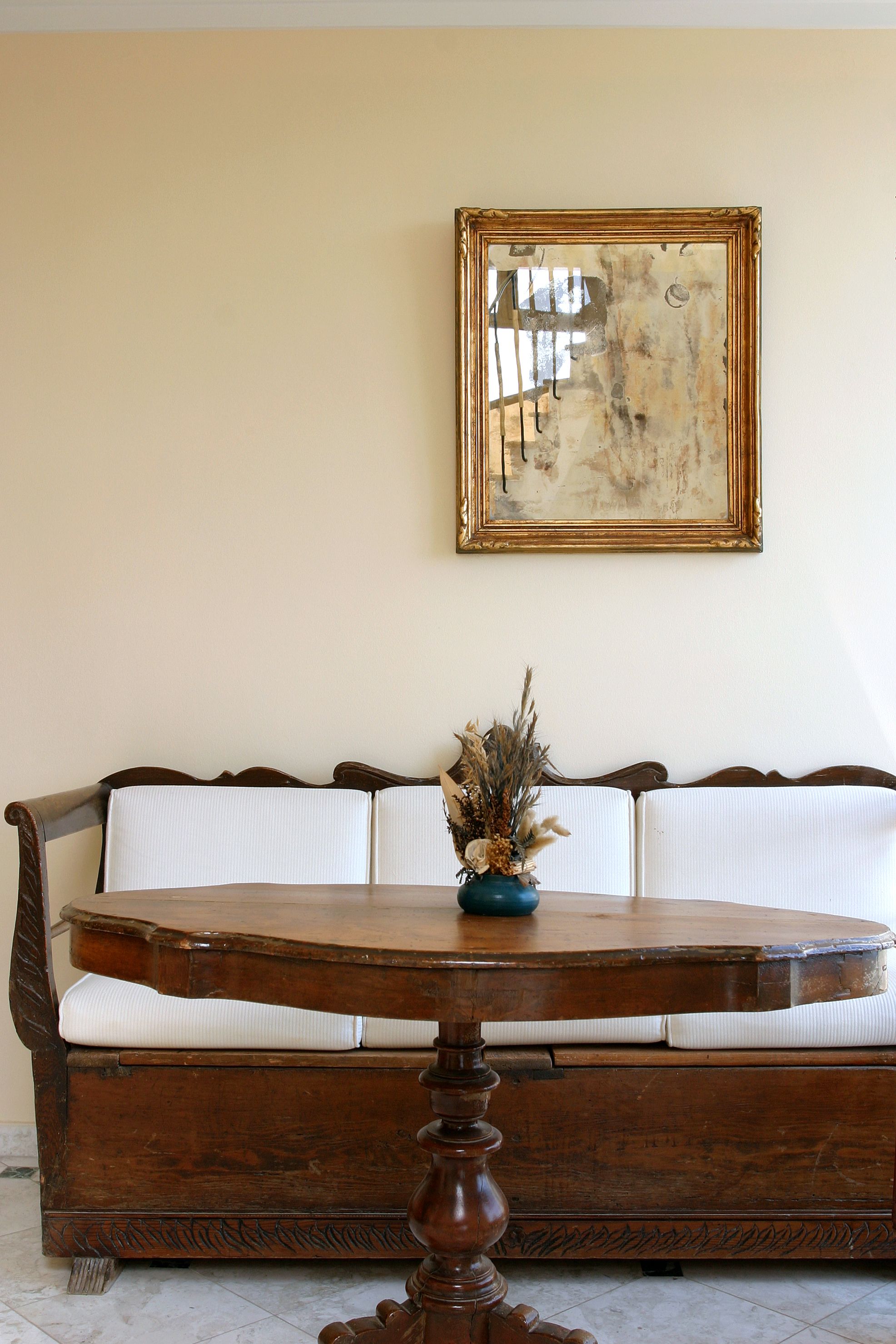 A wooden table with a vase of dried flowers, a wooden bench with white cushions, and a framed abstract painting on the wall.