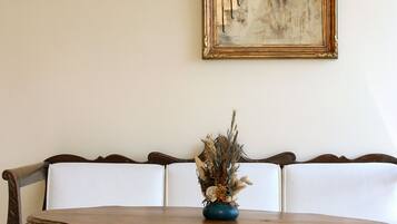 A wooden table with a vase of dried flowers, a wooden bench with white cushions, and a framed abstract painting on the wall.
