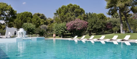 A pool with clear blue water, surrounded by white lounge chairs and a small white building.