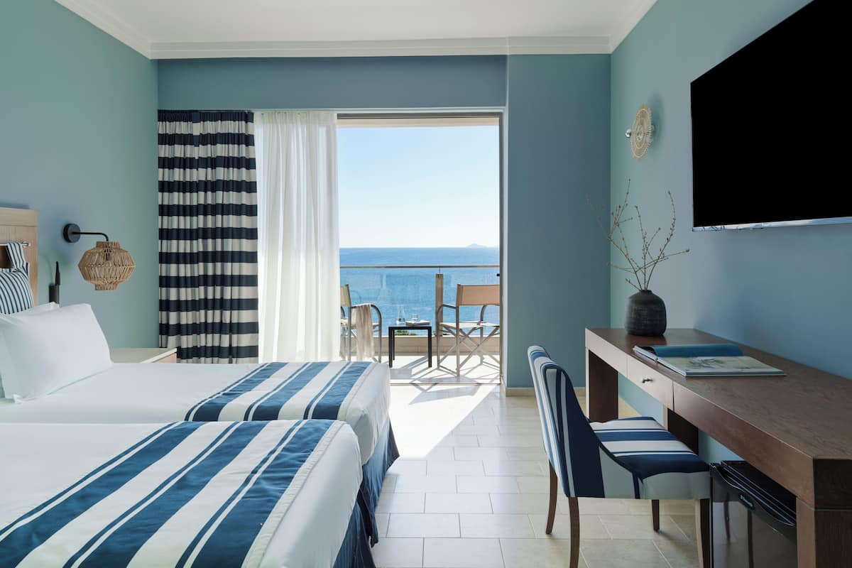 A hotel room with two beds, a desk, a chair, a TV, and a view of the sea.