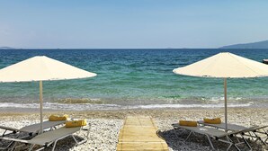 Une plage avec deux parasols et des chaises longues, une promenade en bois menant à l’eau, et une vue dégagée sur la mer et la côte lointaine.