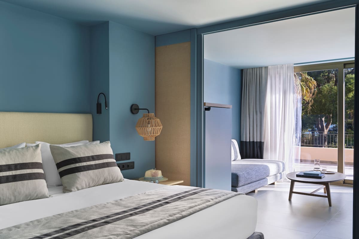 A modern hotel room with a large bed, a bedside table, a lamp, and a view of the outdoors through a sliding glass door.