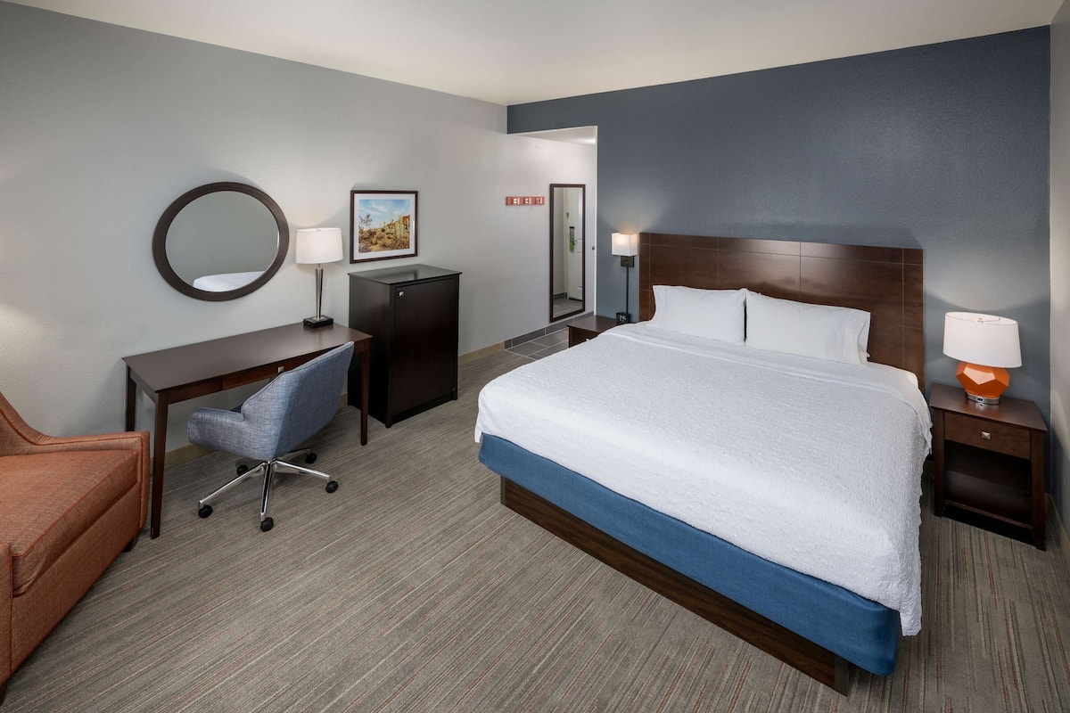 A hotel room with a large bed, a desk with a chair, a small cabinet, a round mirror, and two bedside lamps.