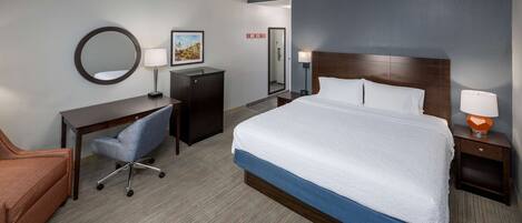A hotel room with a large bed, a desk with a chair, a small cabinet, a round mirror, and two bedside lamps.