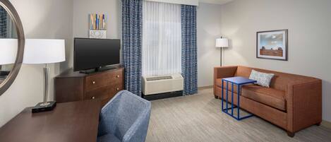 Ett hotellrum med ett skrivbord i trä, en tv, en blå stol, en brun soffa, ett litet bord, en lampa, ett fönster med gardiner och en inramad tavla på väggen.