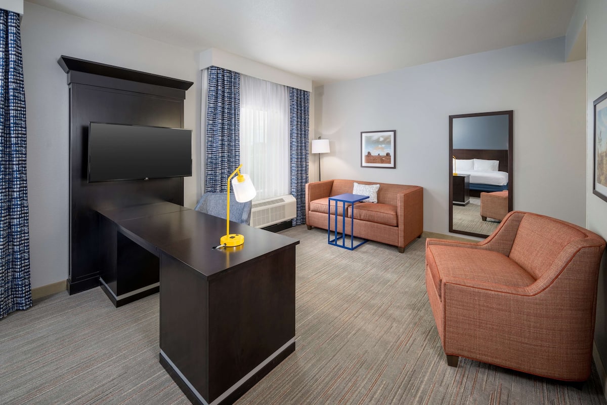 A hotel room with a flat-screen TV, a desk with a lamp, a sofa, a chair, and a bed.