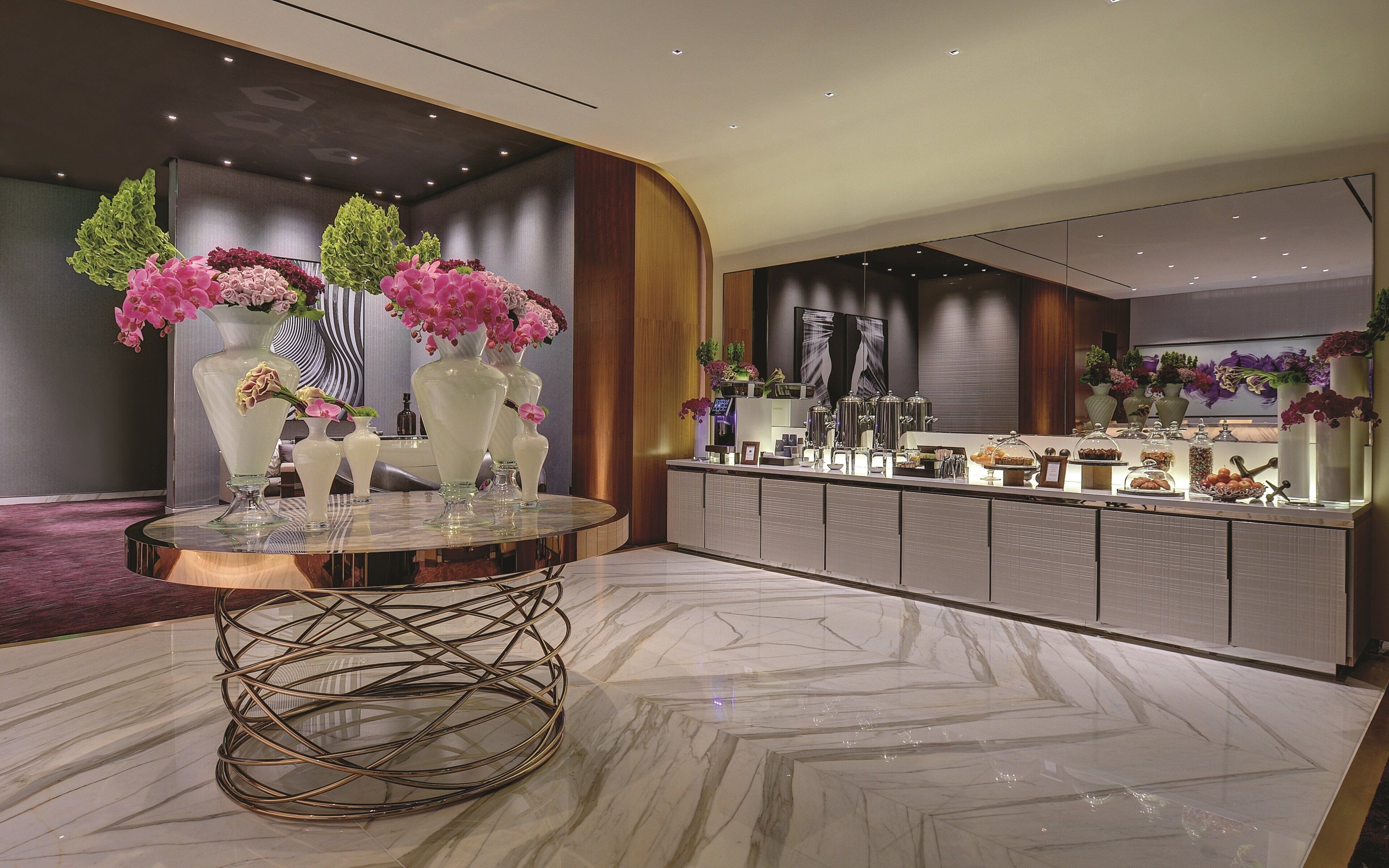 Una hall d'hotel moderna con pavimento in marmo, un tavolo rotondo con vasi e un'area buffet sullo sfondo.
