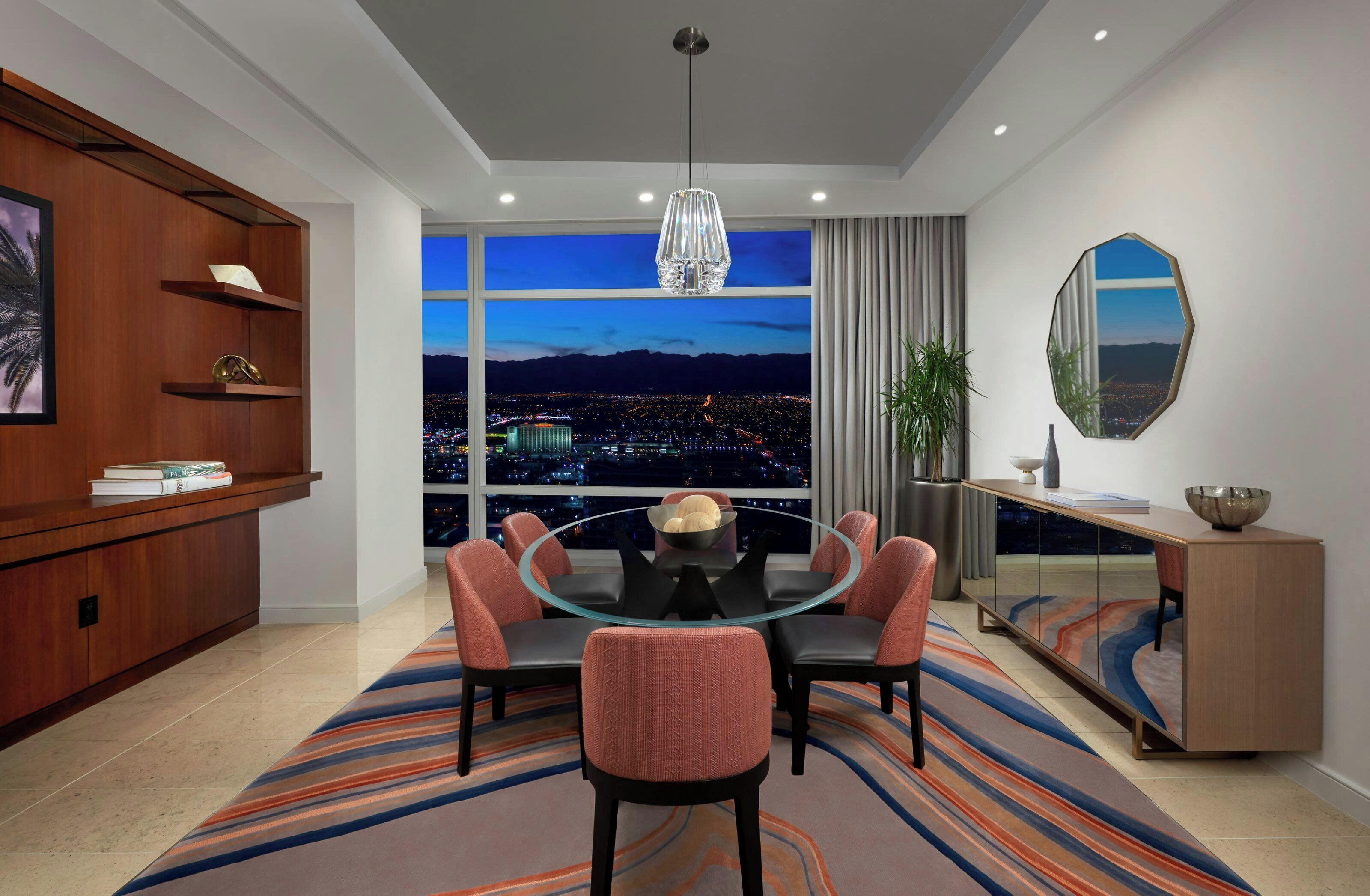 A modern dining room with a round glass table, six chairs, a large window with a city view, a wooden cabinet with shelves, and a decorative mirror.