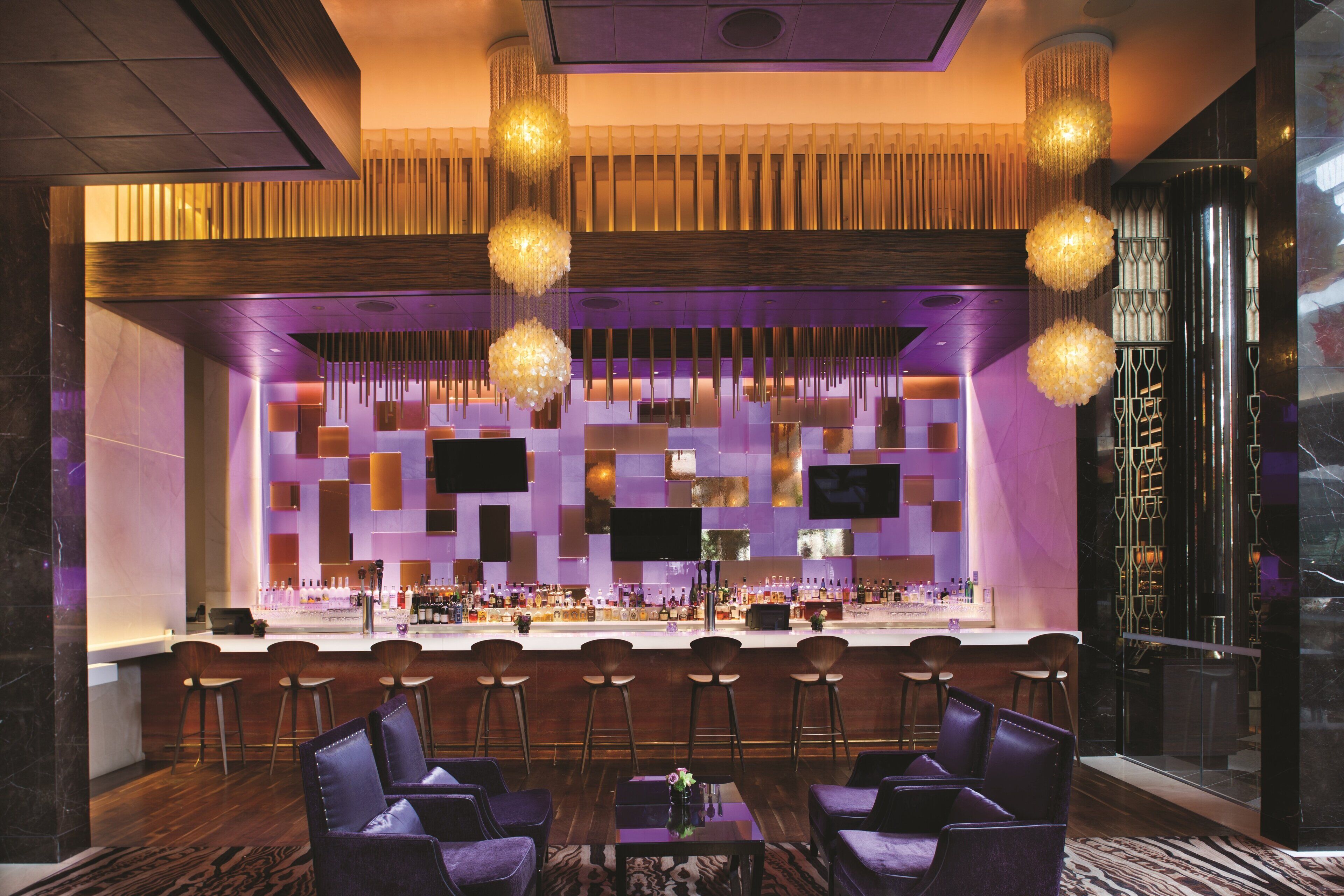 Un espace bar au design moderne, comprenant un long comptoir avec des tabourets et un coin salon avec des fauteuils recouverts d’un tissu violet.