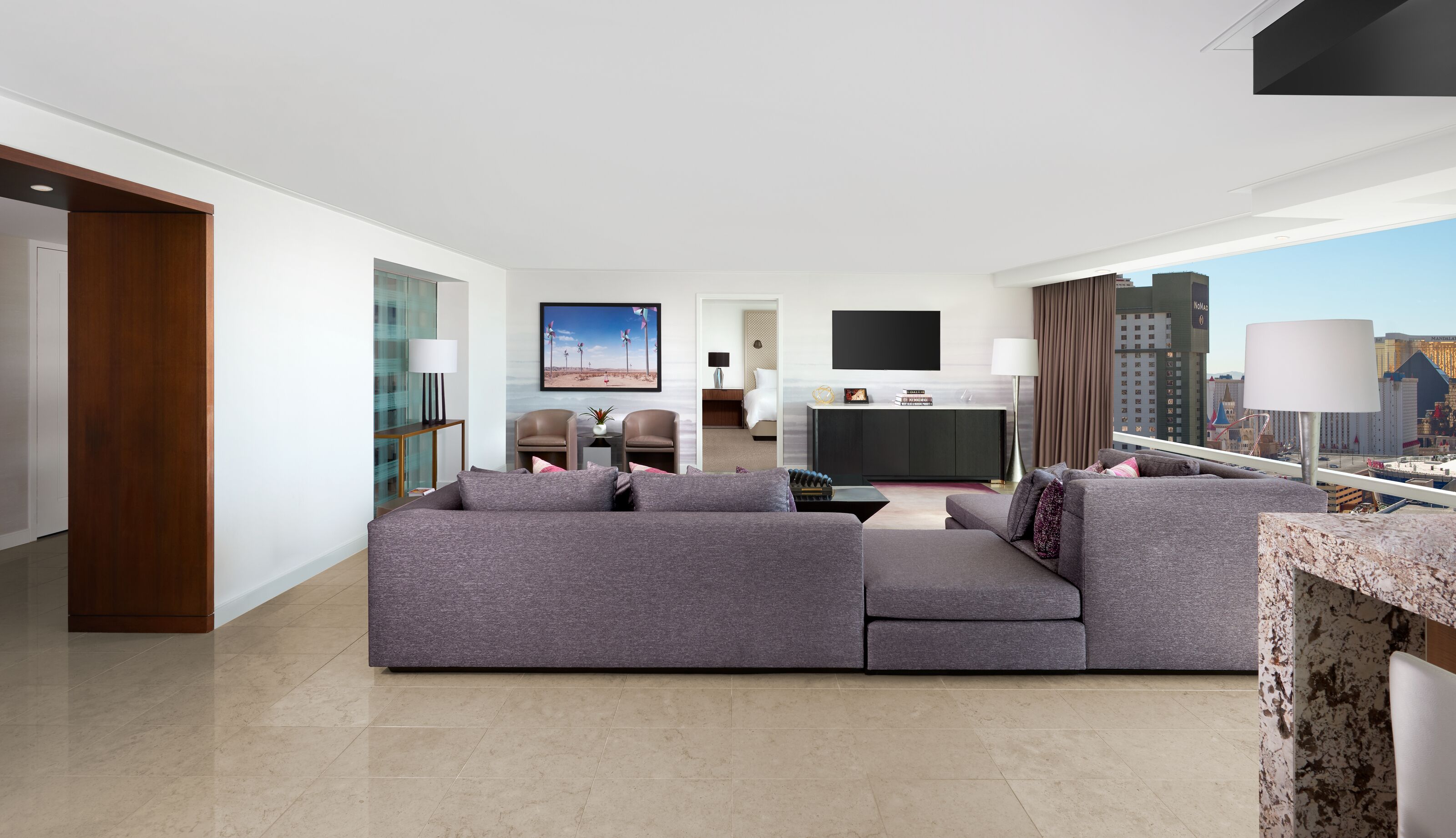 A modern hotel room with a large gray sectional sofa, a flat-screen TV, a desk with a lamp, and a city view.
