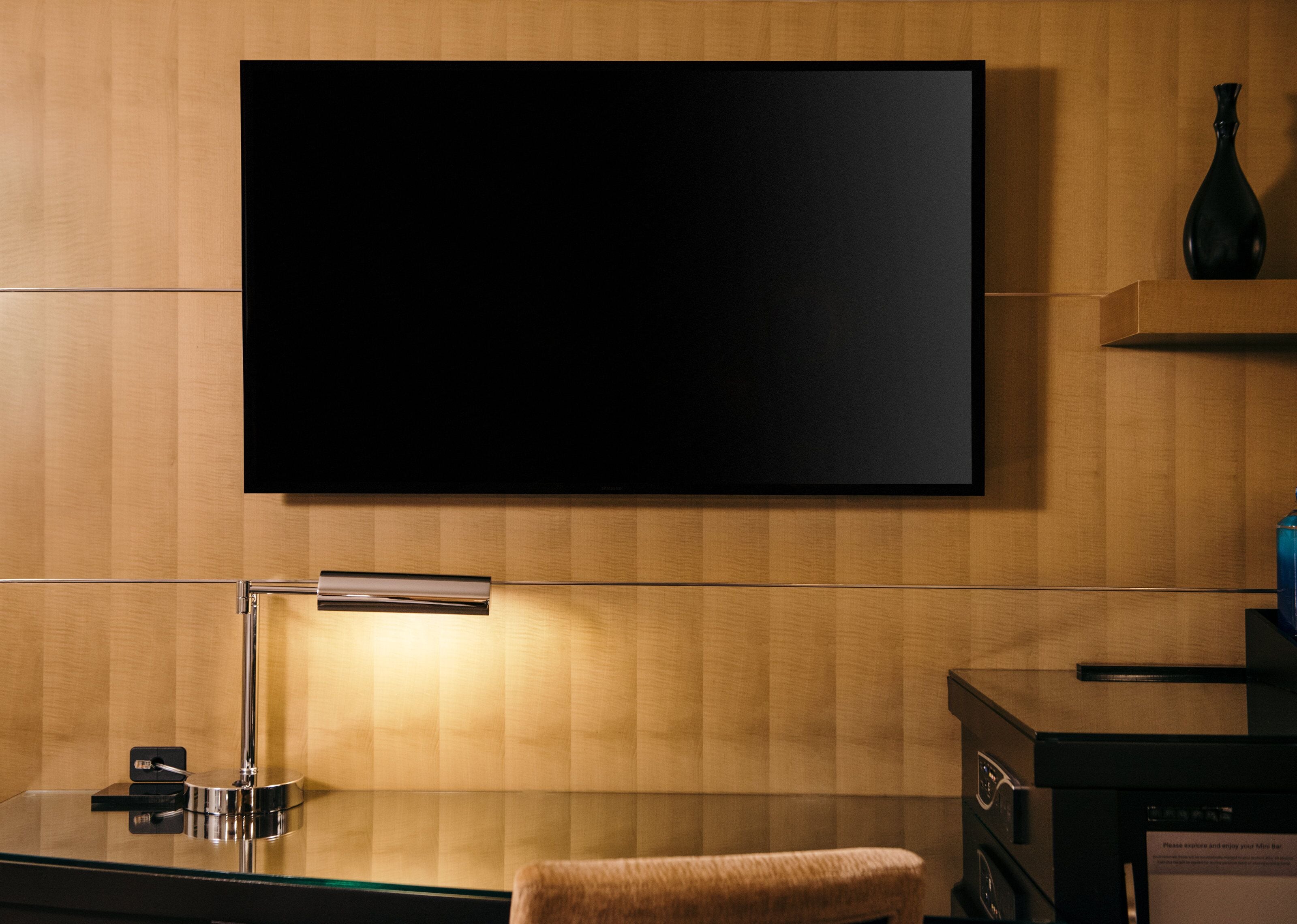A modern room with a flat-screen TV mounted on the wall, a desk with a lamp, and a small seating area.