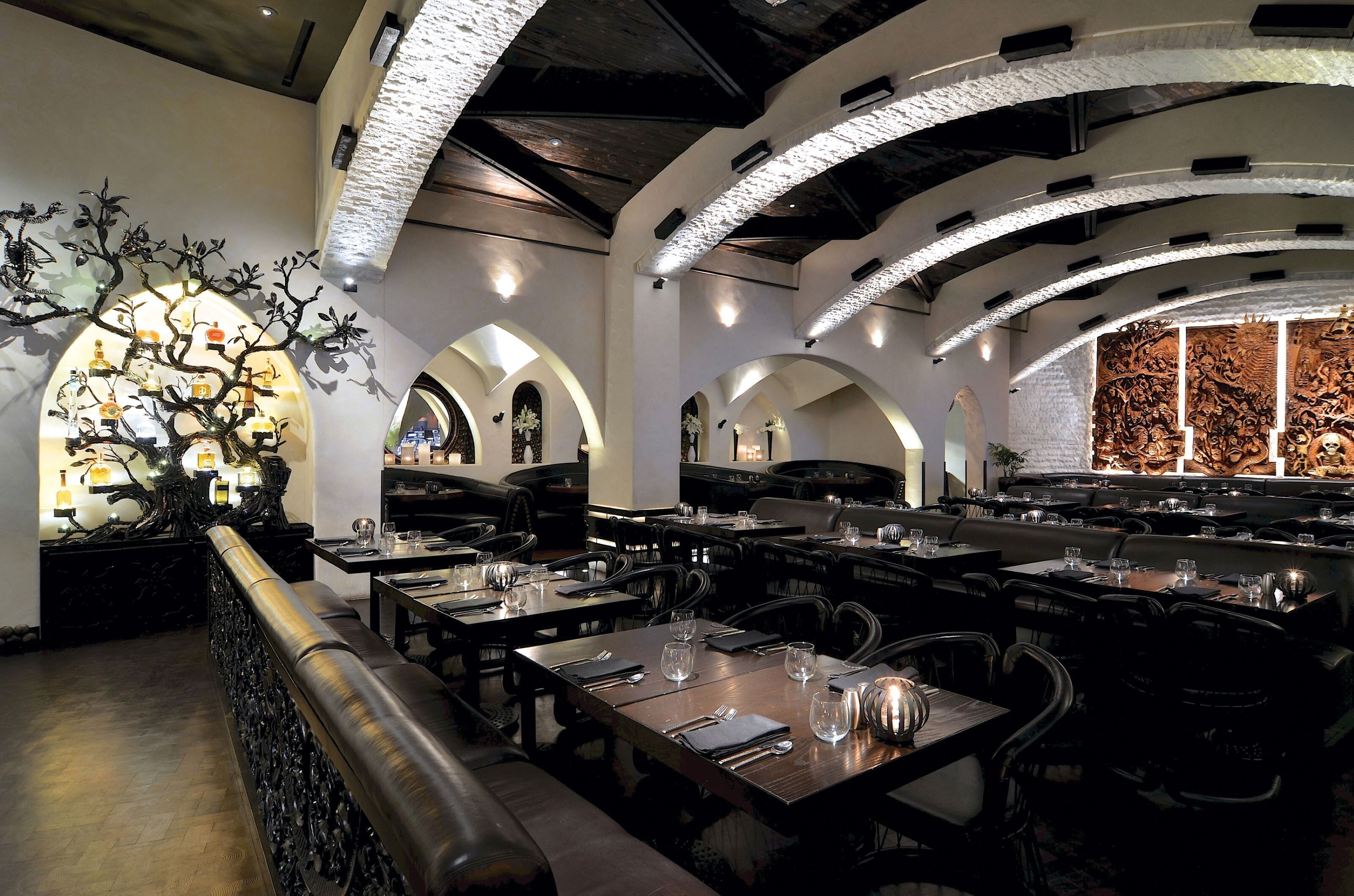 Un restaurant au décor moderne, avec des plafonds en forme d’arc et une sculpture d’arbre décorative au centre. Les tables sont dressées avec des verres à vin et des couverts.