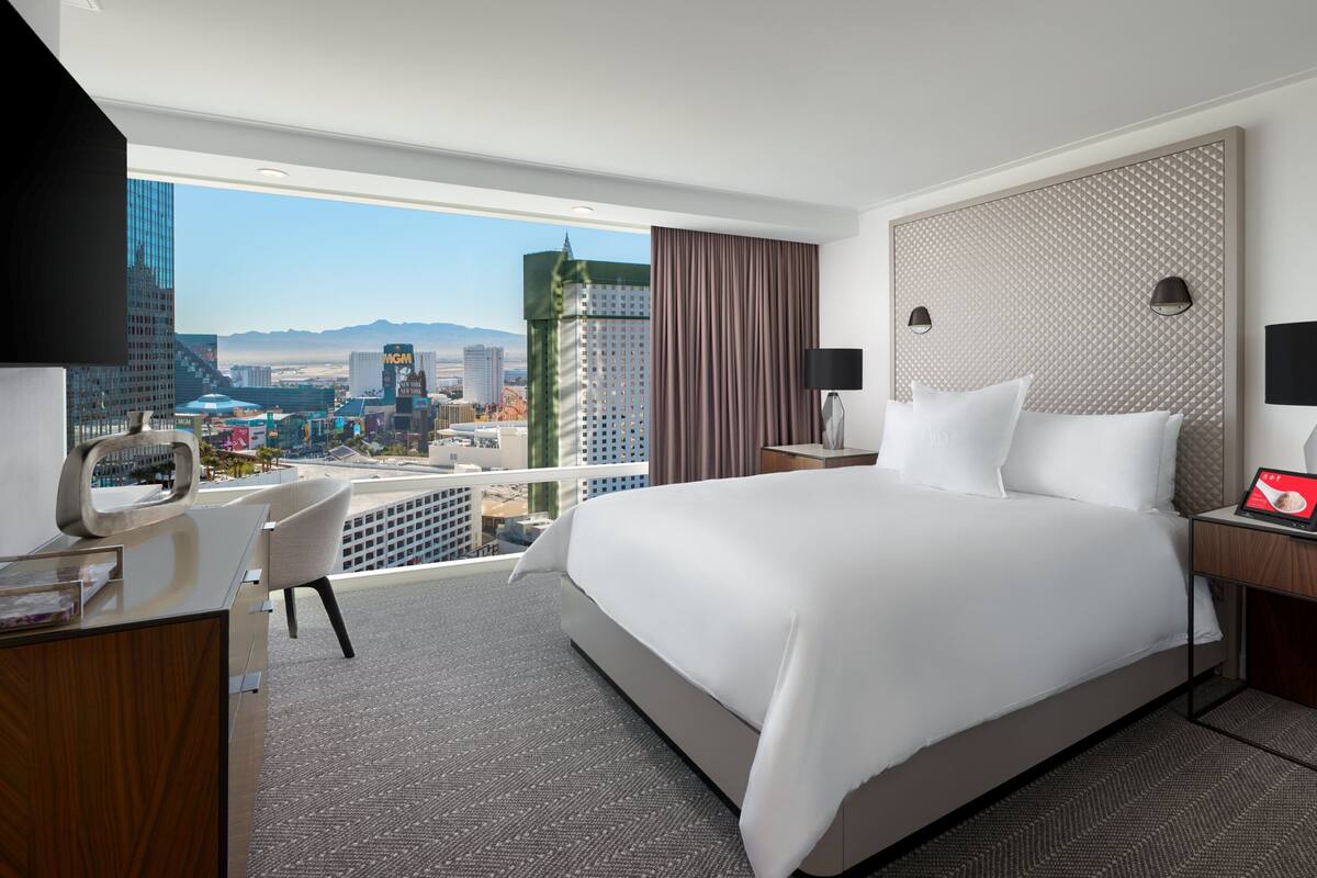 Ein Hotelzimmer mit einem großen Bett, einem Schreibtisch, einem Stuhl, einem Fernseher und Blick auf eine Stadtlandschaft mit Gebäuden und Bergen.