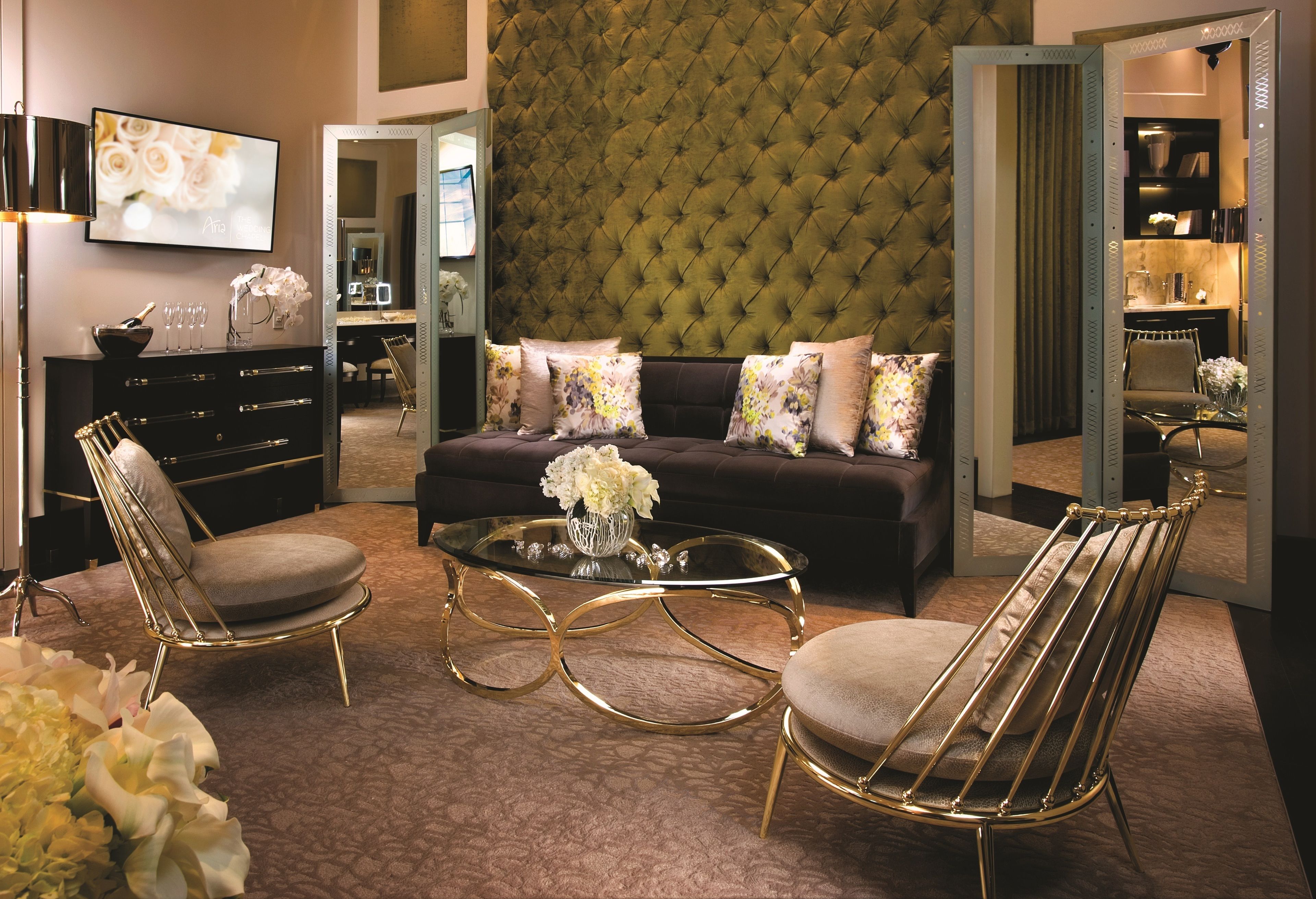 A living room with a tufted green wall, a dark sofa with floral cushions, a glass coffee table, and two gold-framed chairs.