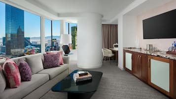 A hotel room with a sofa, a coffee table, a TV, a desk, and a view of the cityscape.