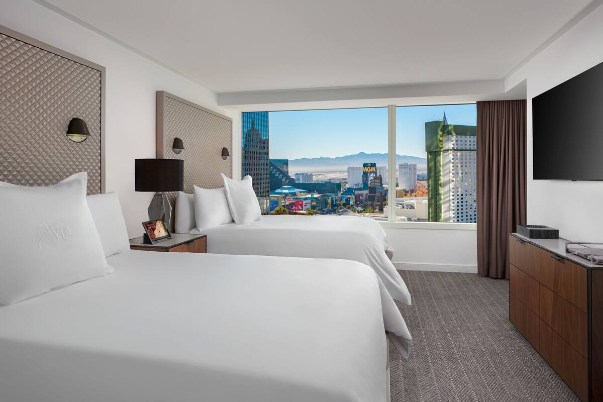 Ein Hotelzimmer mit zwei Betten, einem großen Fenster mit Blick auf die Stadt, einem Flachbildfernseher und einer Holzkombination.