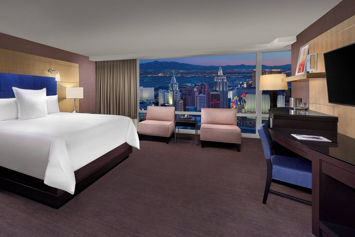A hotel room with a large bed, two armchairs, a desk, and a TV. The room has a city view.