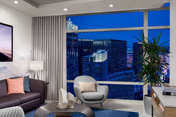 Een moderne woonkamer met uitzicht over de stad, een bank, een fauteuil, een bijzettafeltje en een potplant.