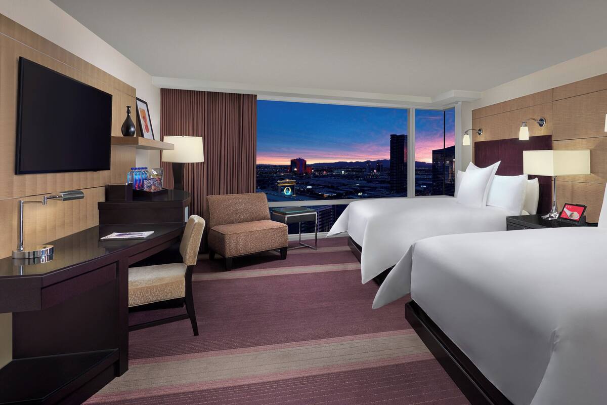 A hotel room with two beds, a desk, a chair, a TV, and a view of a cityscape at dusk.