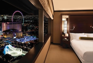 Ein Hotelzimmer mit Bett, Nachttisch, Lampe und Blick auf eine belebte Stadtlandschaft bei Nacht, darunter ein Riesenrad und ein Casino.