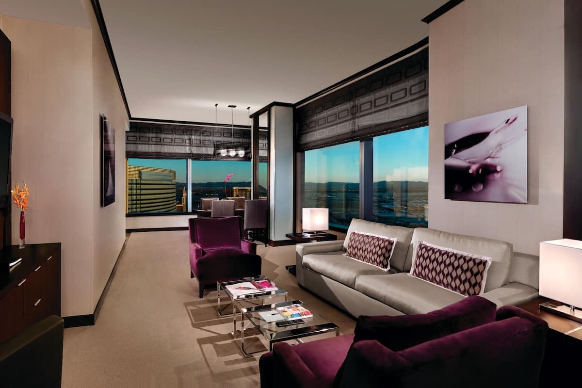 Habitación de hotel moderna con un ventanal amplio que ofrece vistas a la ciudad, un sofá, una silla, una mesa de centro y un televisor.
