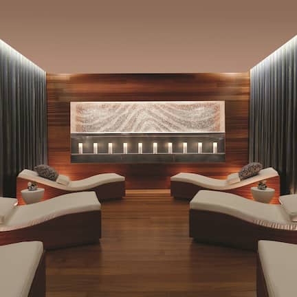A spa room with two reclining chairs, a wooden wall panel, a decorative wall piece, and a small table with a plant.